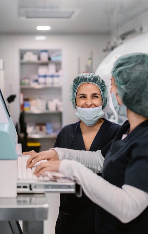 Two Women Biologists Working On Microscope In IVF Laboratory, smiling at each other and typing into a system. Our practice area of medical device and healthcare.